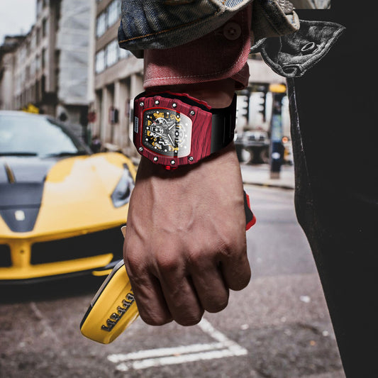 Hand wearing a red and black watch with a blurred city street and yellow car in the background